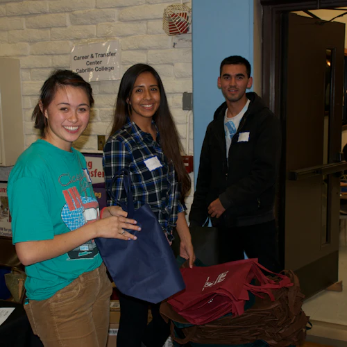 three smiling students in casual dress standing in the gym in front of a brick wall