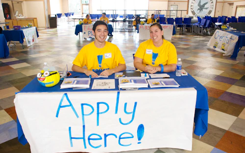 estudiante sentado en una mesa con un letrero de "SOLICITA AQUÍ"