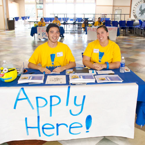 student sitting at a table with an "APPLY HERE sign