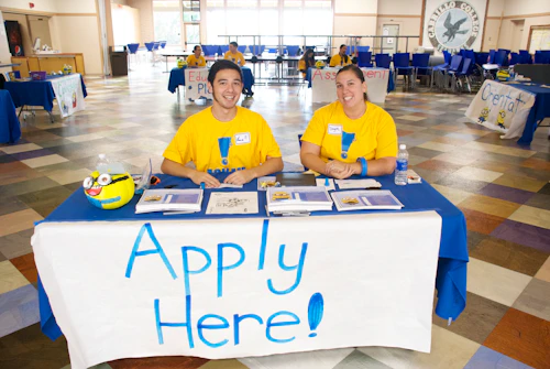 student sitting at a table with an "APPLY HERE sign