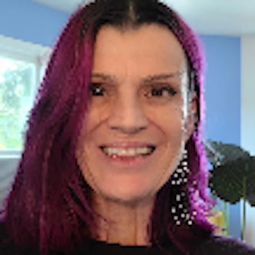 Sadie smiling in a blue room with plants; she has light skin and purplish hair, blue wall,