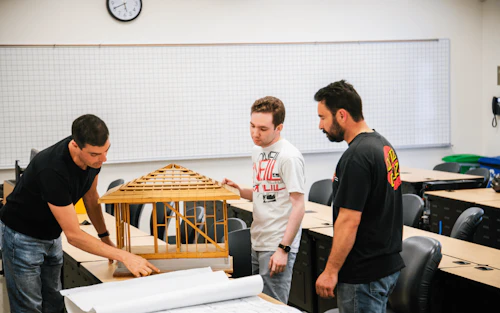 Estudiantes de arquitectura mirando planos en un escritorio mientras uno sostiene una maqueta de madera de una casa.
