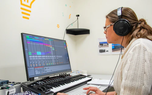Una mujer con auriculares grandes está sentada frente a una computadora con un teclado musical debajo de la pantalla.