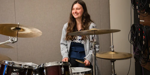 A smiling woman sits on a stool playing a set of drums.
