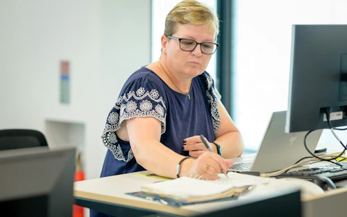 Mujer sentada en una computadora y escribiendo en un bloc de notas sobre un escritorio.