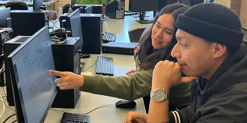 Estudiante mujer señalando el monitor de la computadora para mostrarle algo al estudiante hombre sentado a su lado.