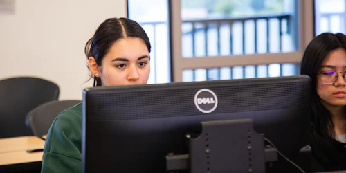 Dos estudiantes mujeres se sientan frente a monitores de computadora mirando atentamente las pantallas.