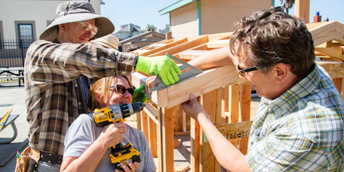 Una mujer perfora un agujero en el marco de una casa mientras dos hombres la ayudan.