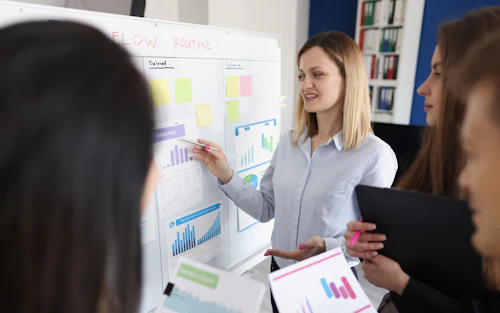 Una instructora señala un gráfico de barras en una pizarra blanca mientras tres estudiantes toman apuntes.