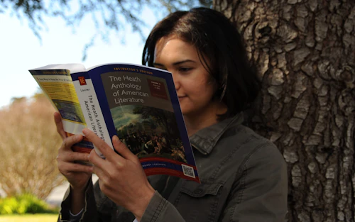 Una estudiante lee un libro mientras se apoya en un árbol.