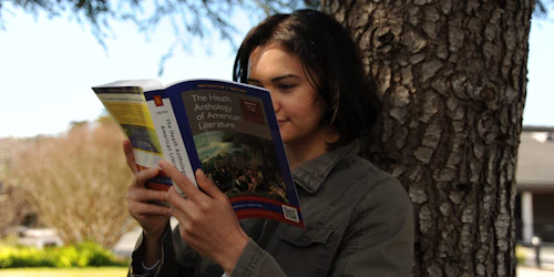 A female student reads a book while leaning on a tree