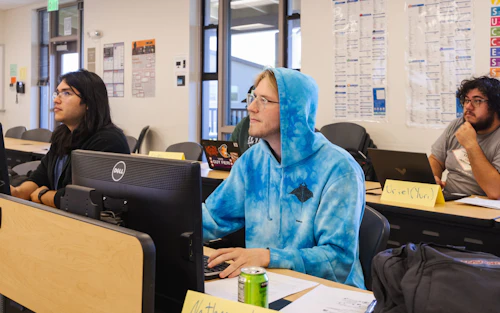 Tres estudiantes varones están sentados en escritorios con monitores de computadora.