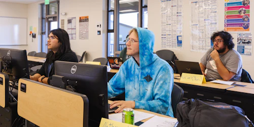 Three male students sit at desks with computer monitors