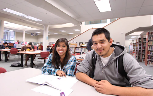 Una estudiante y un estudiante están sentados en un escritorio en una biblioteca.