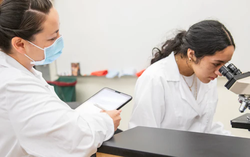 Una estudiante con bata de laboratorio observa a otra estudiante mirando a través de un microscopio.