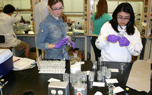Una estudiante con guantes y gafas de seguridad manipula un tubo de ensayo mientras otra estudiante observa.