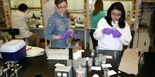 Una estudiante con guantes y gafas de seguridad manipula un tubo de ensayo mientras otra estudiante observa.