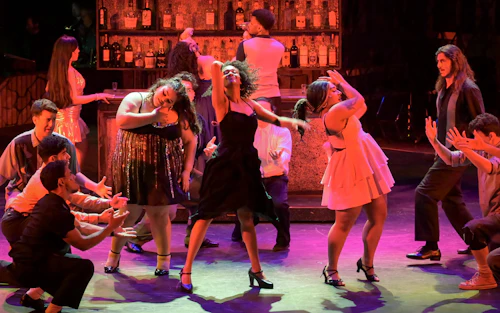 Una mujer canta rodeada de otros actores durante una presentación de In the Heights.