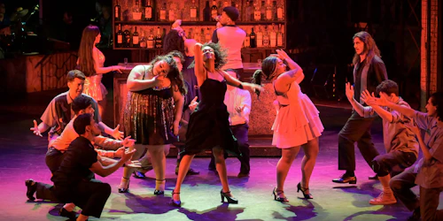 A woman sings surrounded by other actors during a performance of In the Heights - Photo by Jana Marcus