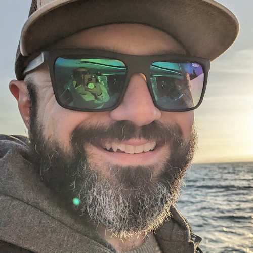 Bearded man on sailboat wearing blue jacket, gray hood, with a hat and sunglasses