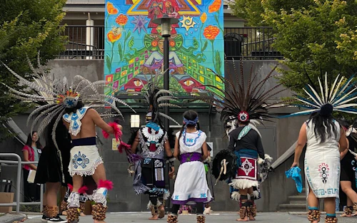 Mitotiliztli dancers, seen from behind, perform on a paved area scattered with flower petals. They wear elaborate feathered headdresses and traditional attire. Behind them is a tall building adorned with a vibrant, colorful mural, flanked by trees.