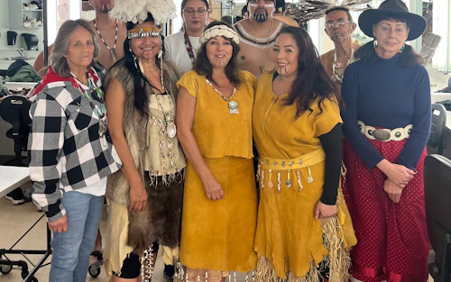 The Amah-ka-tura cultural dance group backstage in the Crocker Theater. Several individuals are dressed in traditional Indigenous regalia, featuring feathered headpieces, shell necklaces, and fringed garments made of what appears to be hide or woven materials.