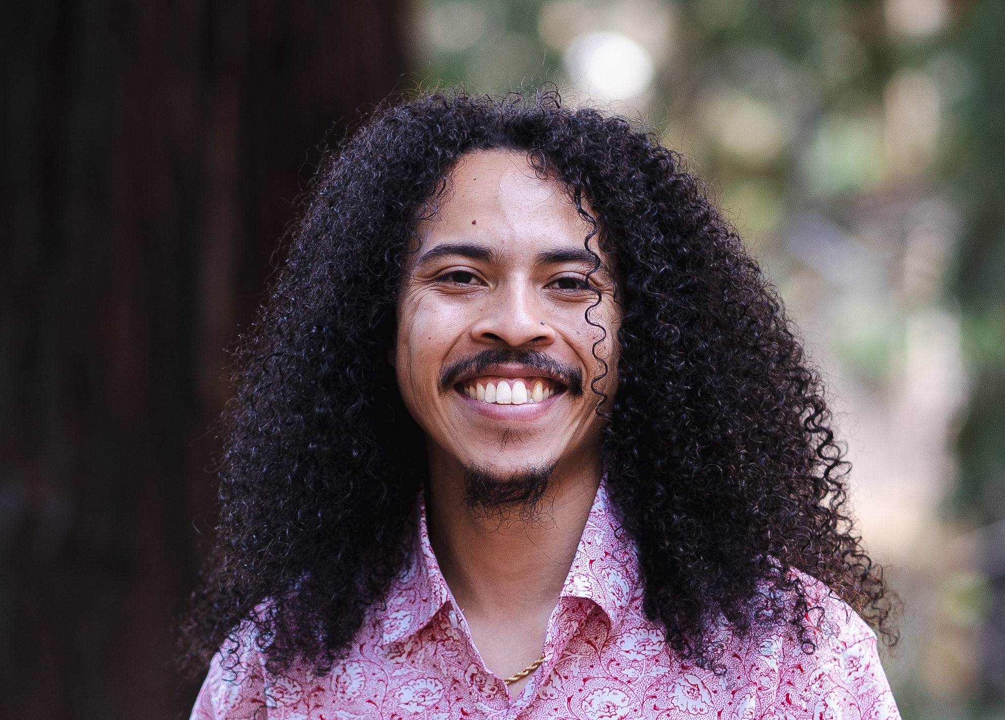 professional headshot of Vincente Perez, a lightskinned multiracial man with long dark brown curly hair. Vincente is smiling and wearing a red and white shirt with blurred redwood trees in the background  