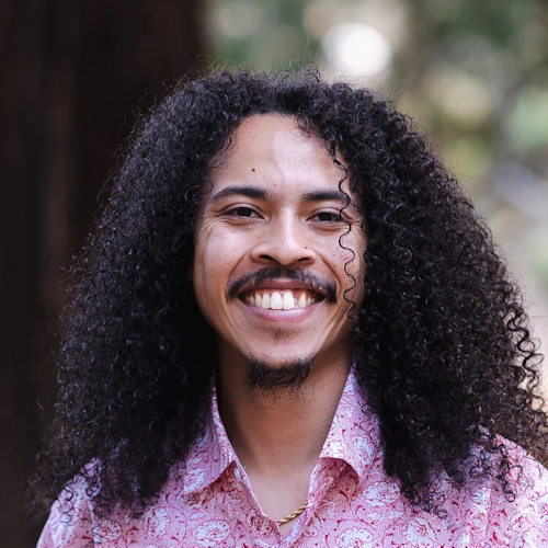 professional headshot of Vincente Perez, a lightskinned multiracial man with long dark brown curly hair. Vincente is smiling and wearing a red and white shirt with blurred redwood trees in the background