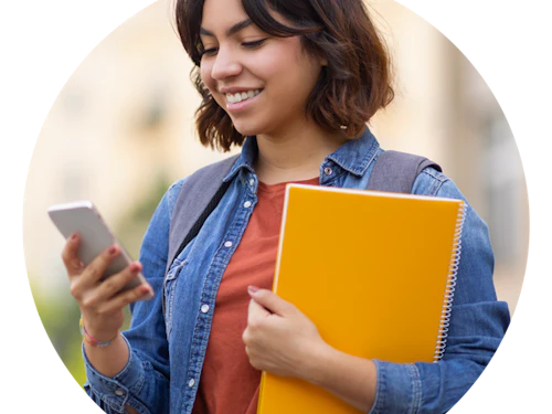 Estudiante universitaria sosteniendo su teléfono y sonriendo a él con una pila de libros en la mano.
