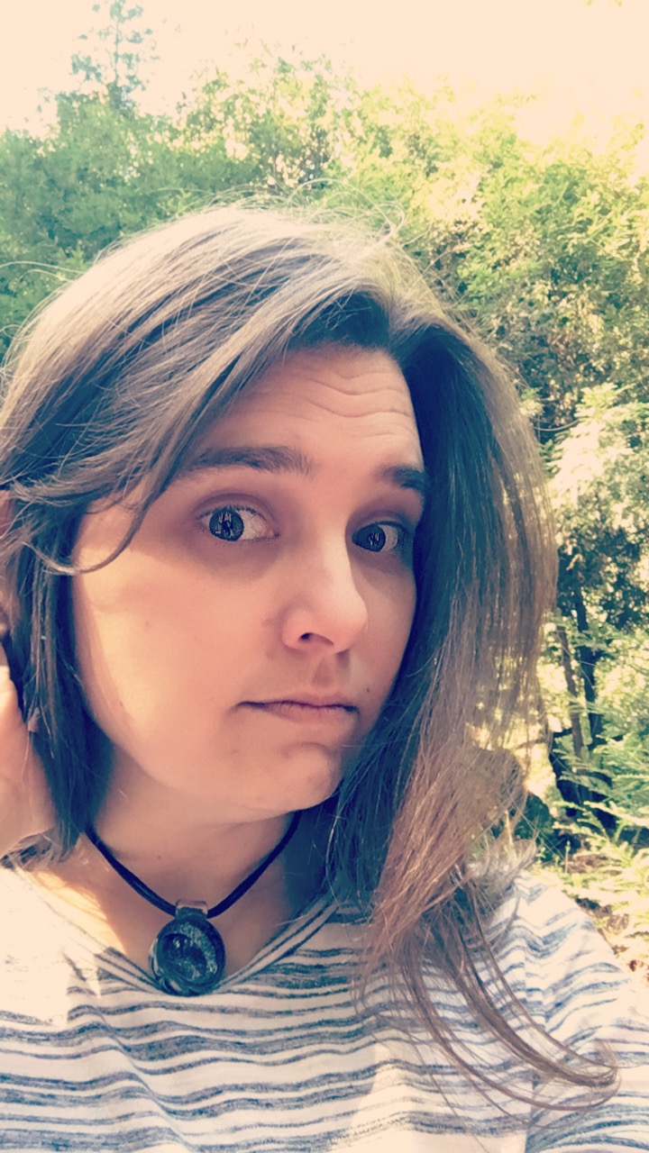 Headshot of Kelly wearing a white and grey stripped shirt, pulling back her medium long brown hair ro the right of her head. She is wearing her famous galaxy necklace standing outside with greenery behind her.