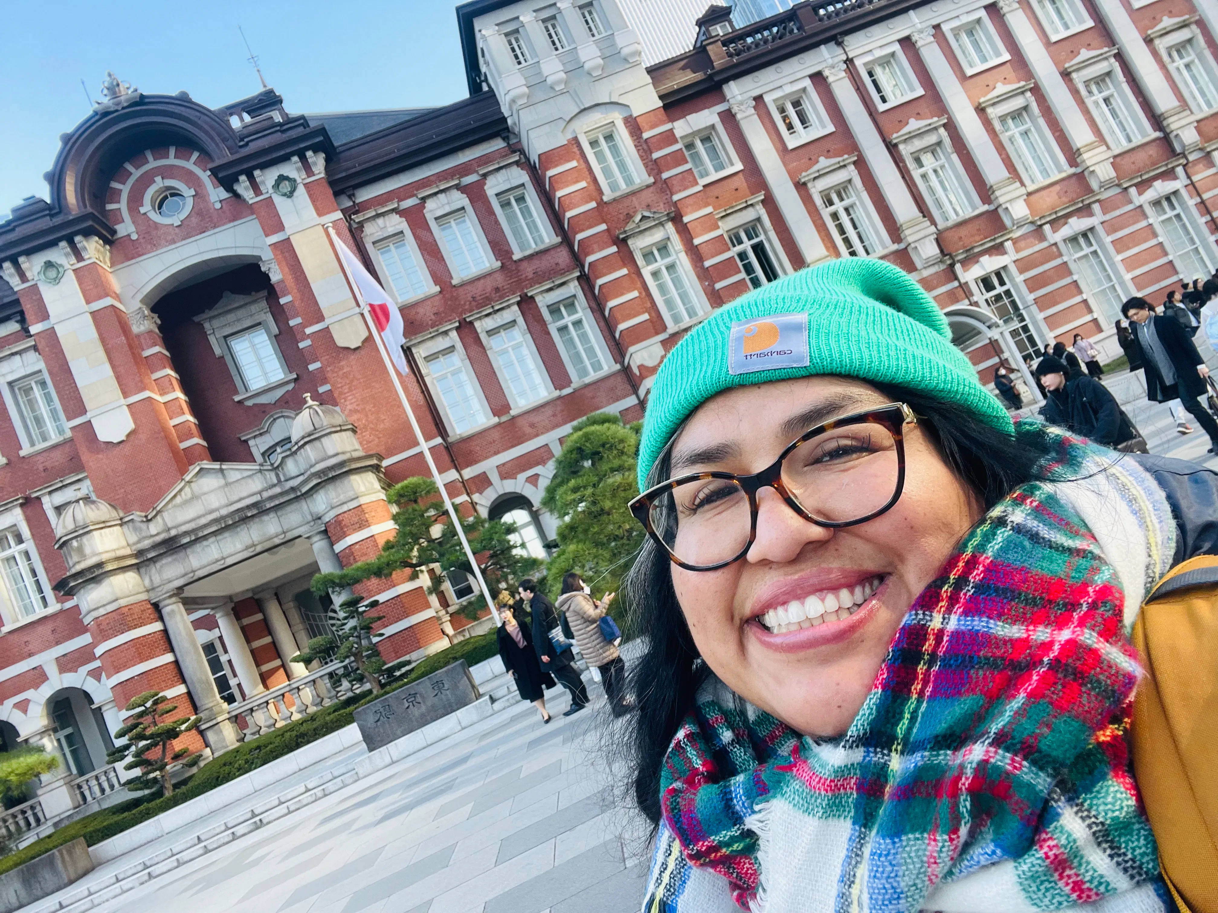 A photograph of Dr. Mogollón wearing a bright teal beanie, plaid scarf, and glasses, standing in front of a brick building in Japan. 