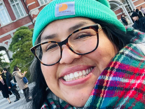 A photograph of Dr. Mogollón wearing a bright teal beanie, plaid scarf, and glasses, standing in front of a brick building in Japan.