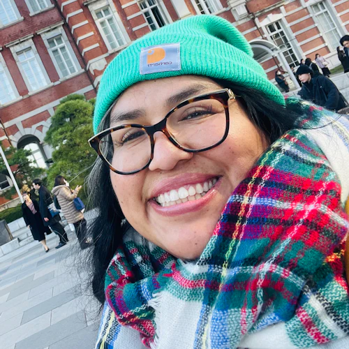 A photograph of Dr. Mogollón wearing a bright teal beanie, plaid scarf, and glasses, standing in front of a brick building in Japan.