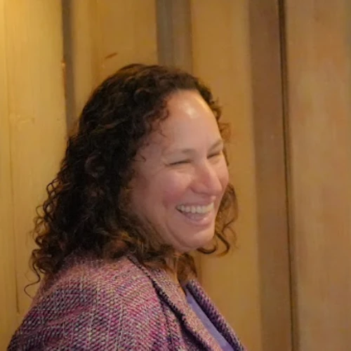 Mixed Race, curly hair Dean smiling in purple top and pink plaid blazer.