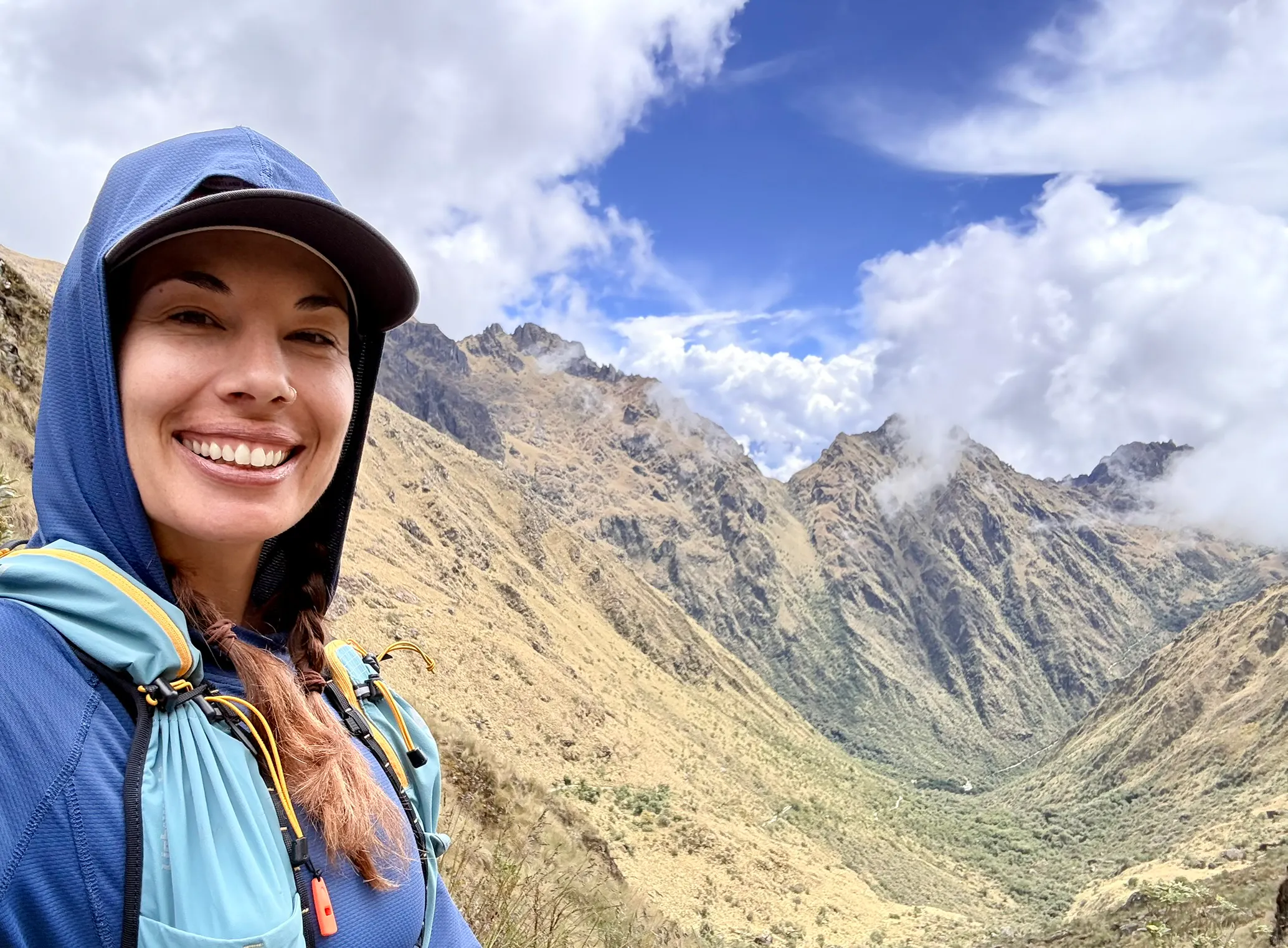 A woman with brown hair and a hood on is shown in front of mountains with clouds in the sky.